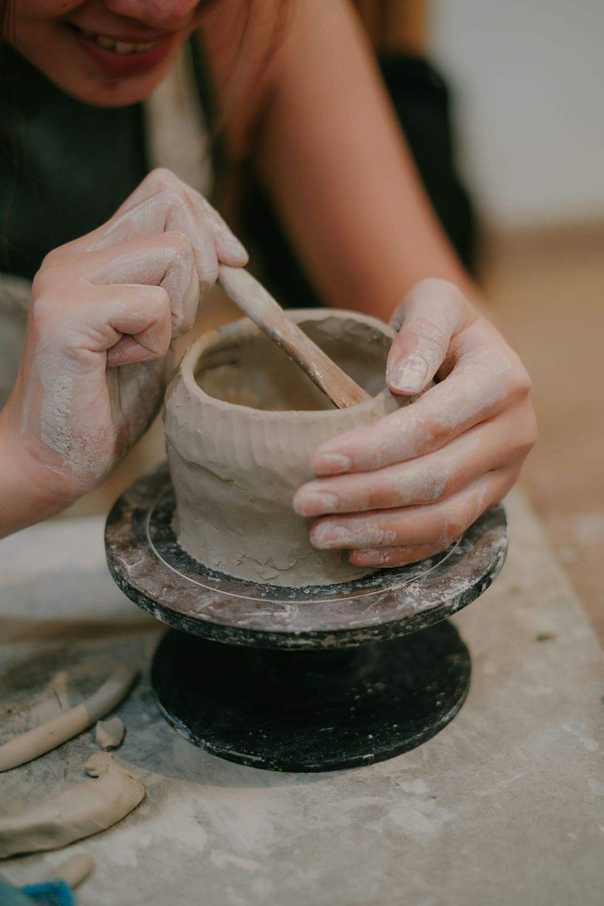 A smiling woman in an apron hand-building a clay pot with a wooden tool, her hands covered in slip, in a warm-lit pottery workshop