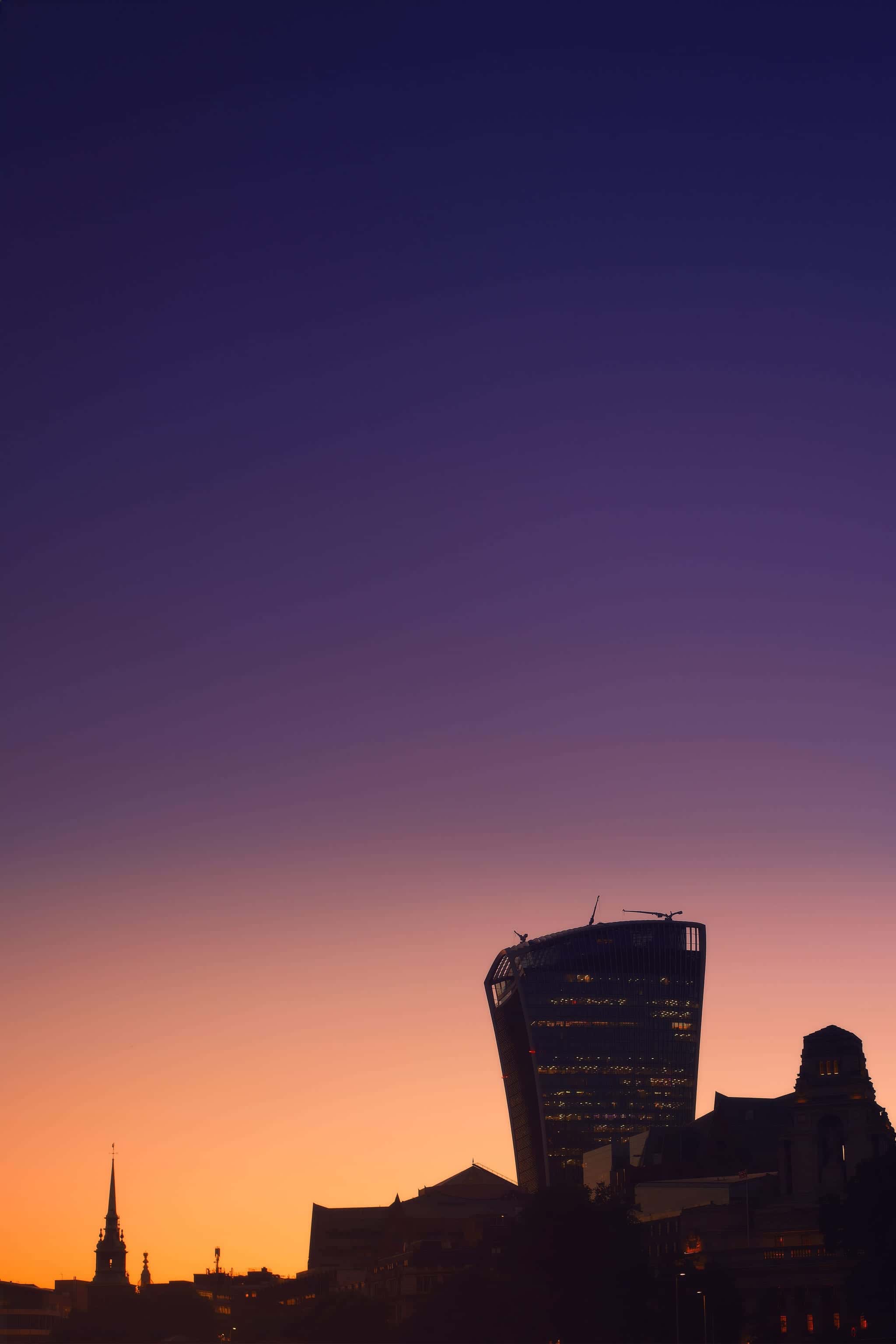 London at twilight — the City of London skyline silhouetted against a deep blue-to-purple night sky that melts into a pink-and-gold sunset glow at the horizon; the rounded Walkie-Talkie tower stands lit on the right, a church spire and riverside buildings on the left